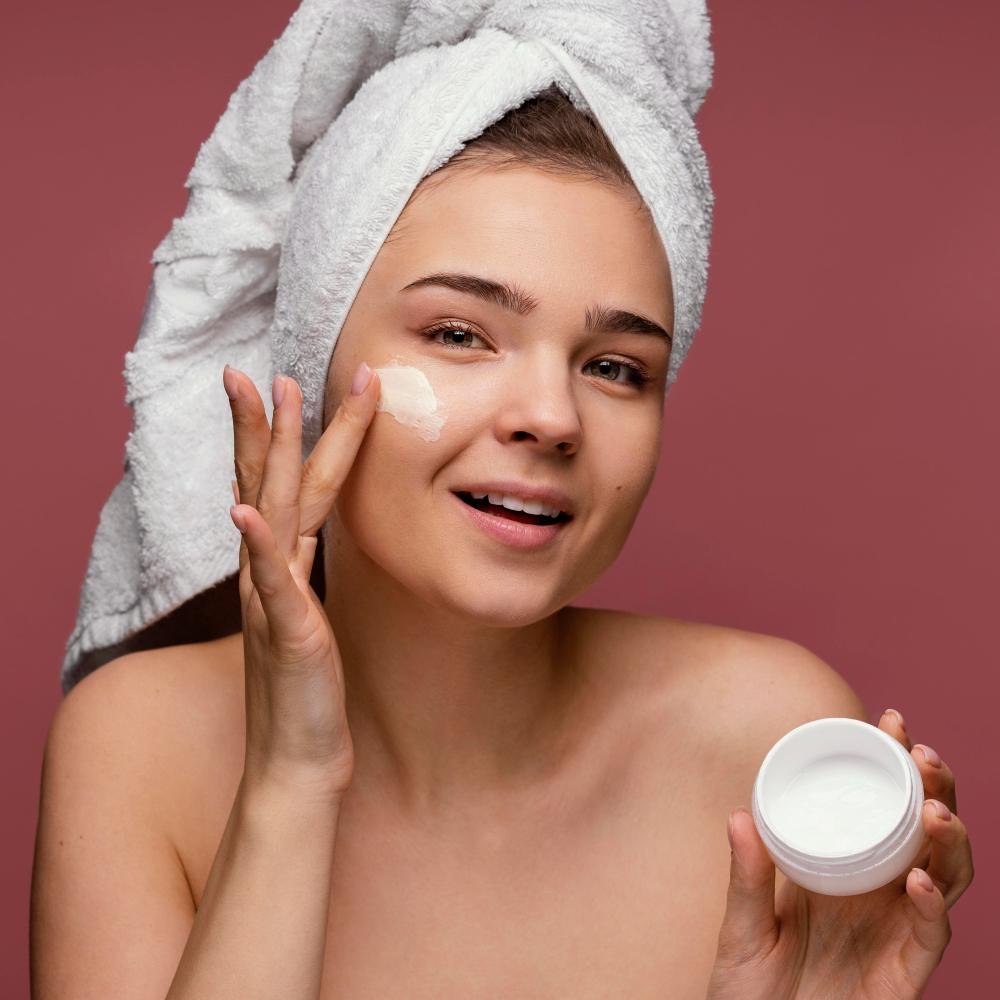 Jeune femme appliquant une crème hydratante sur le visage, serviette sur les cheveux